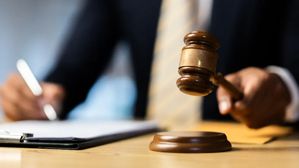Close-up of a wooden judge's gavel held above a sound block, with a person writing on a clipboard in the background.