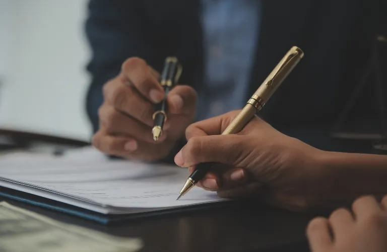 Two individuals holding gold and black pens over a document on a dark surface.